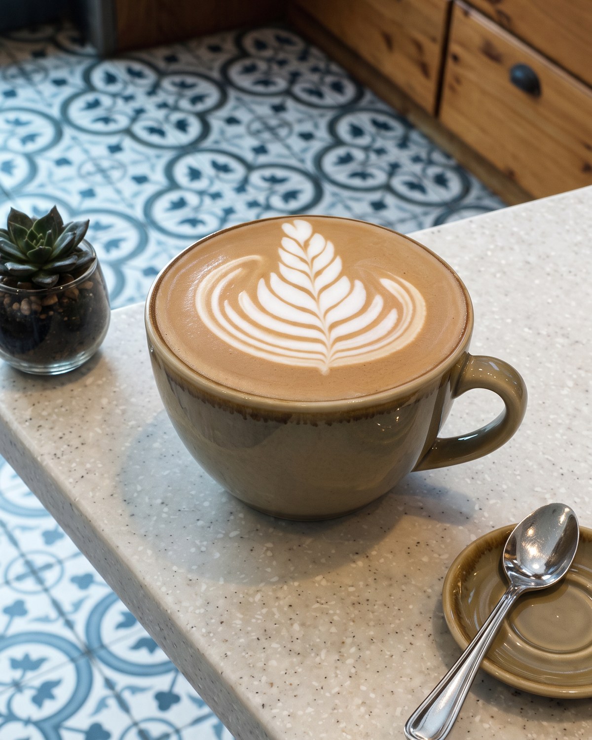 A flat white with fern latte art in a green glazed cup on the terrazzo counter, with the café’s patterned tiles behind