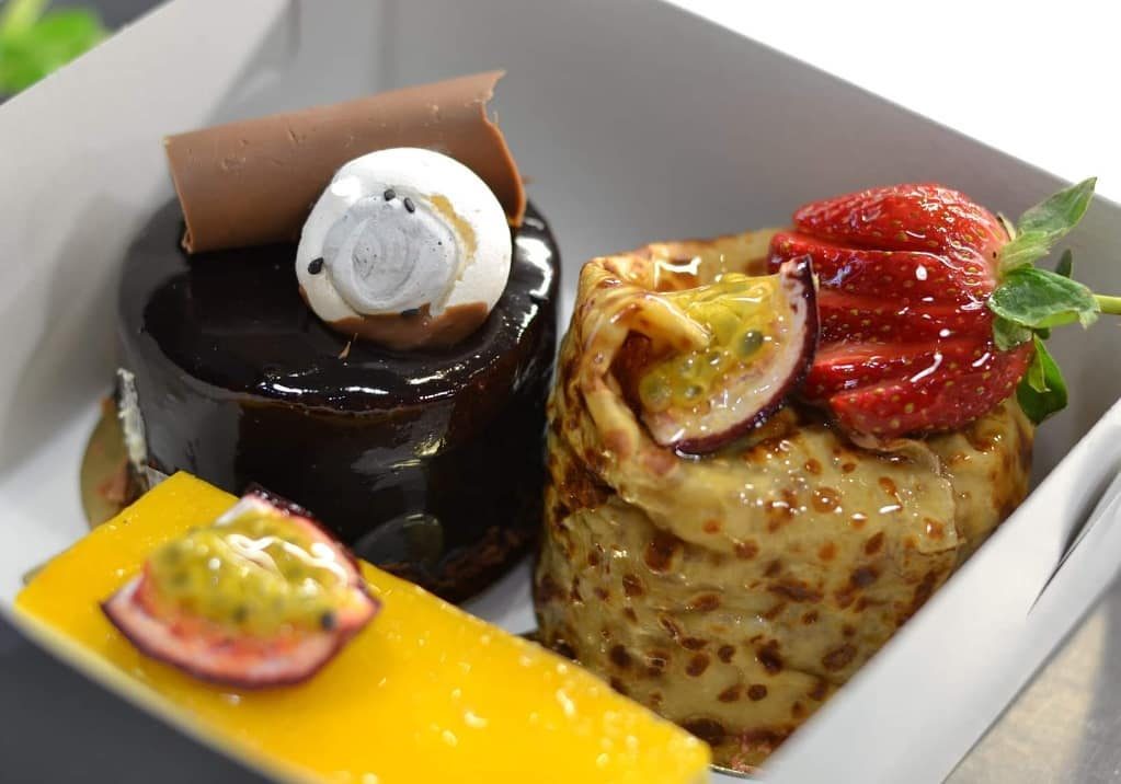 A takeaway box holding a chocolate entremet, mango slice and crêpe cake with strawberry